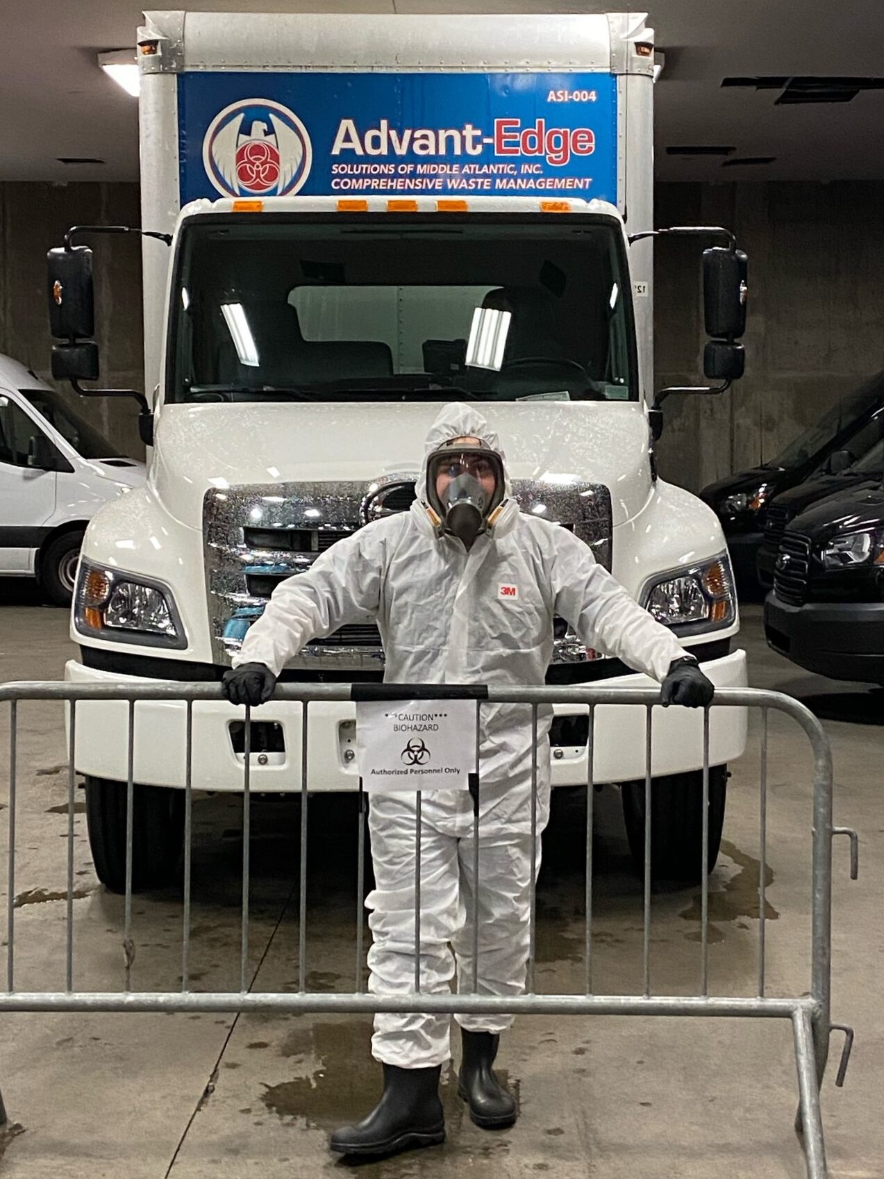 Person in a hazmat suit stands behind a biohazard barricade in front of a truck for hazardous waste disposal.