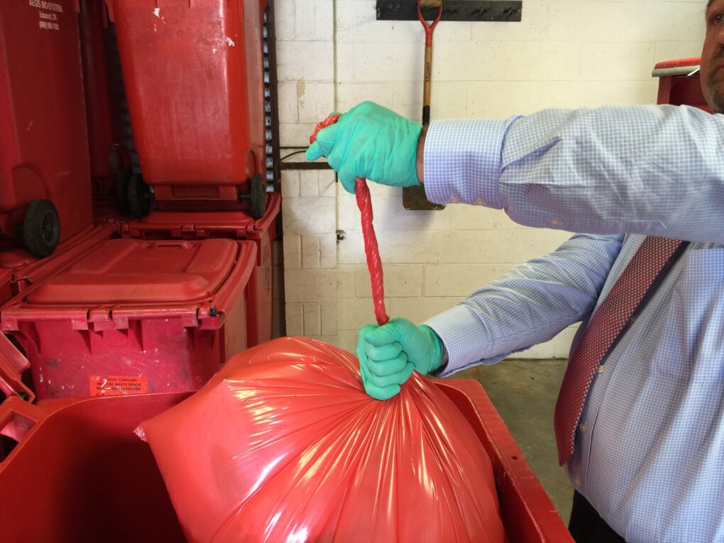 Gloved worker secures red bag for disposal into medical waste containers and packaging bins.